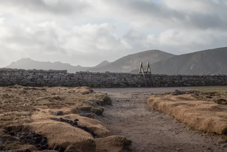 Chemin de terre menant à un muret en pierres avec échelle, montagnes brumeuses à l’arrière-plan.